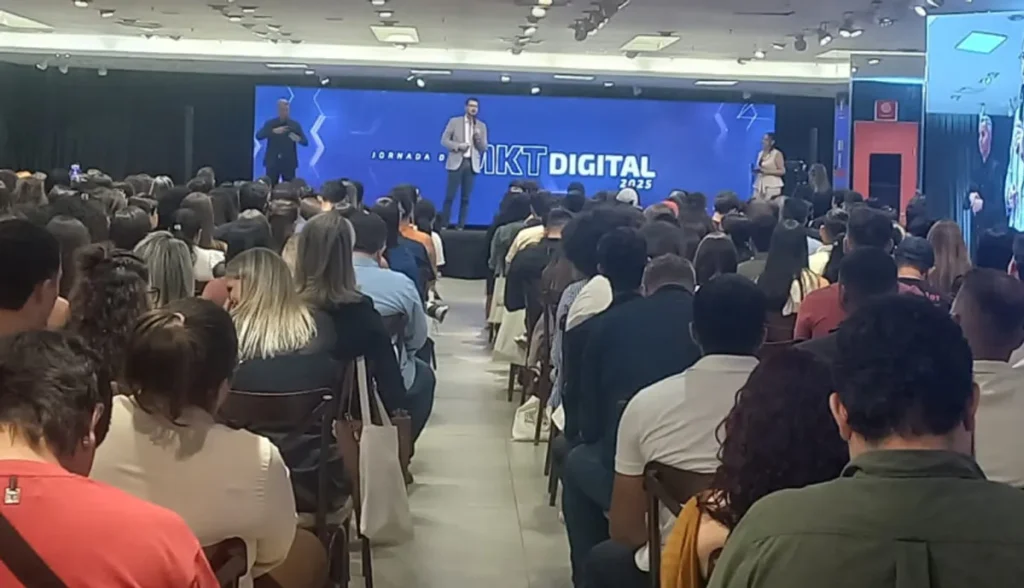 Palestrantes renomados no palco da Jornada de Marketing Digital em Cuiabá, com plateia engajada e telão com o tema do evento e logo do Sebrae/MT.